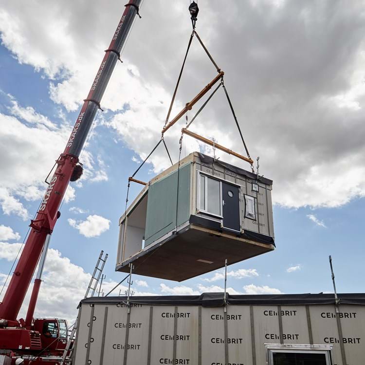 Præfabrikerede moduler løftes sikkert på plads som del af en moderne og bæredygtig byggeproces. “Montage af modulbyggeri – kran løfter præfabrikeret bygningsmodul under sikre og kontrollerede forhold.”